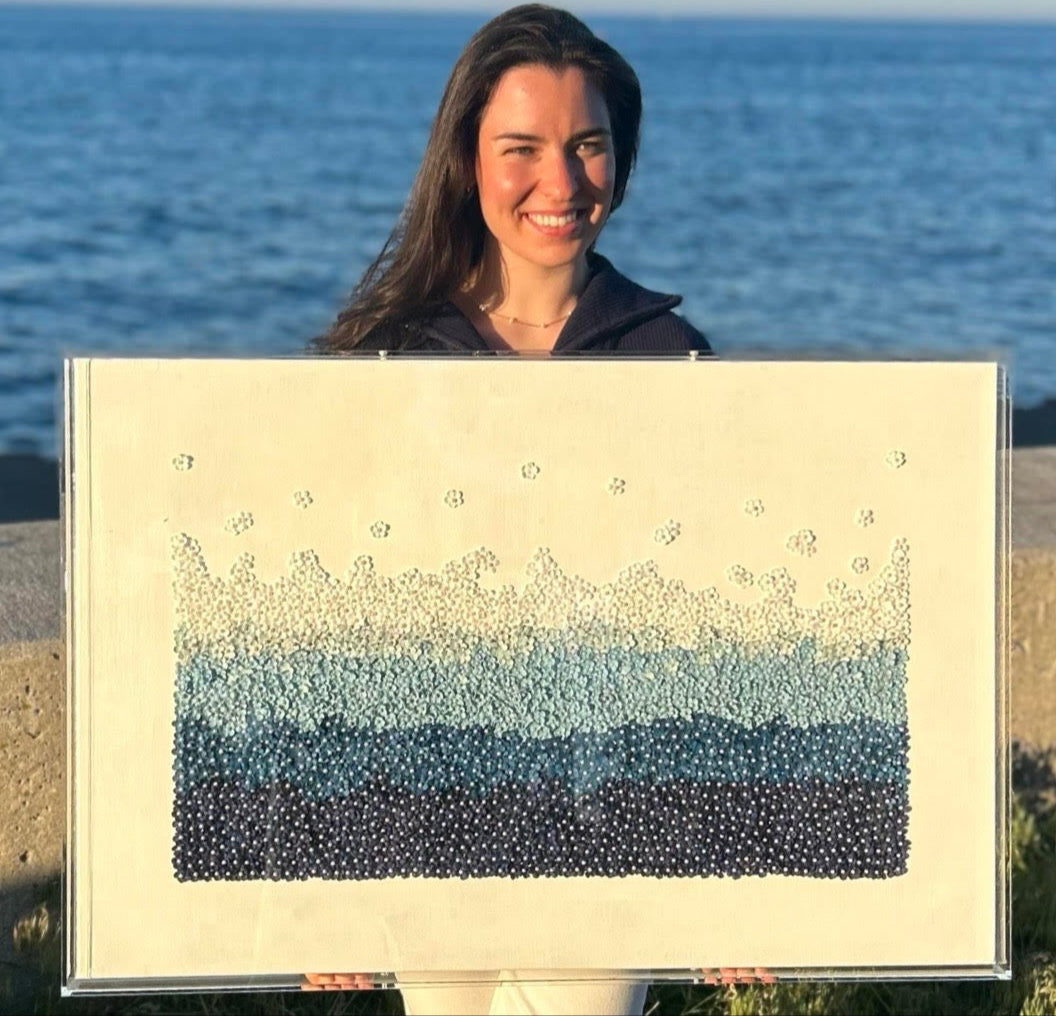 A person standing by a body of water holding a framed artwork with a floral design made from dyed mulberry paper flowers and imitation pearls.